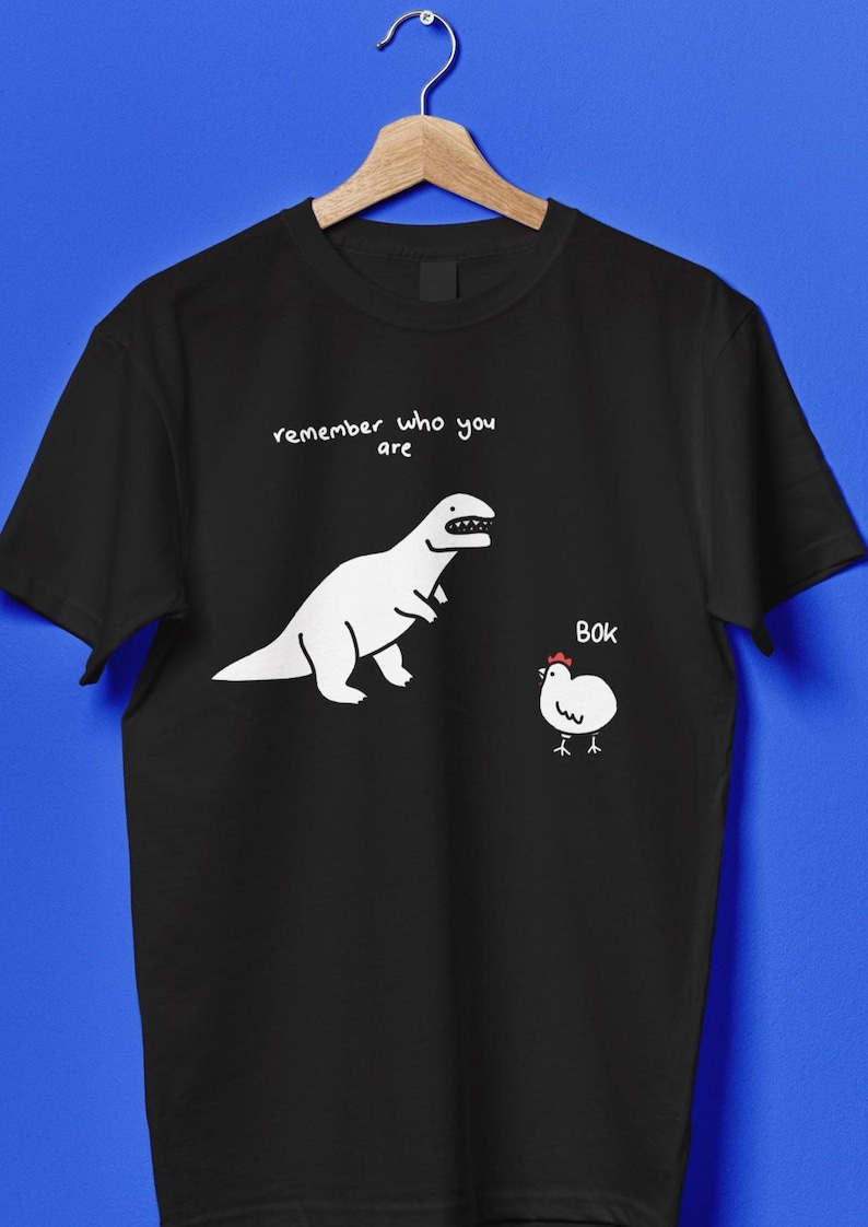 t-shirt with a dinosaur and chicken graphic. The text "remember who you are" is above the dinosaur. The chicken has the text "BOK" next to it.