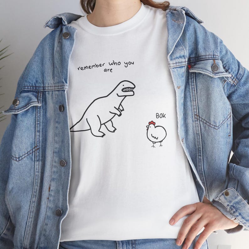 May include: A white t-shirt featuring a black line drawing of a dinosaur and a chicken. The text "remember who you are" is printed above the dinosaur, and the word "Bok" is printed above the chicken, which has a small red comb on its head.