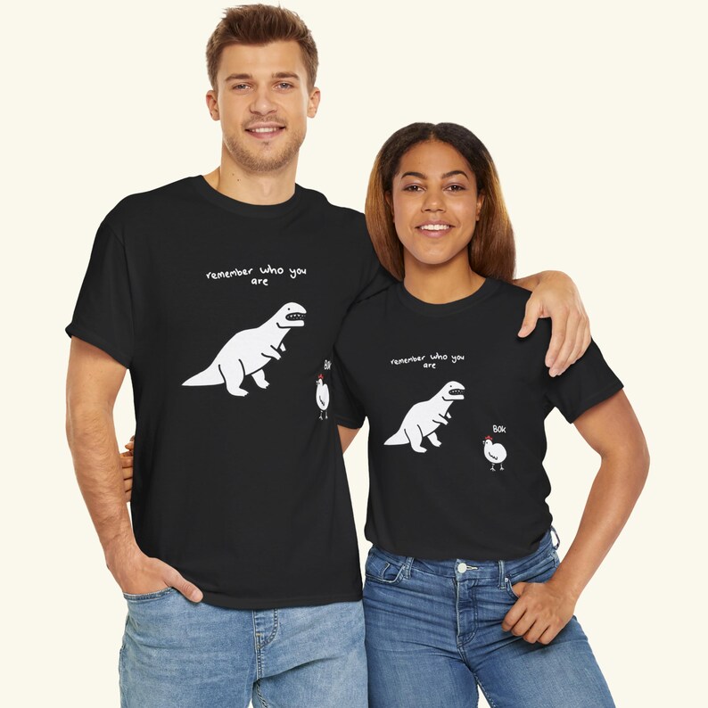 May include: A black short-sleeved t-shirt featuring a white graphic of a dinosaur looking at a chicken. Above the dinosaur is the text "remember who you are" and next to the chicken is the text "Bok". The design is printed on the front of the shirt.