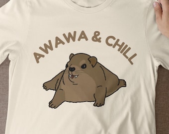 Könnte beinhalten: Cremefarbenes T-Shirt mit einem braunen Cartoon-Tier und dem Text "AWAWA & CHILL" in Braun. Das Tier hat einen lächelnden Ausdruck. Das Shirt ist aus einem weichen Material.