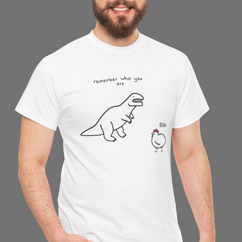 May include: A white short-sleeved t-shirt featuring a black line drawing of a dinosaur and a chicken. Above the dinosaur, the text reads "remember who you are" and next to the chicken, the text reads "BOK". The chicken has a small red comb.