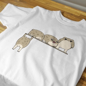 May include: White t-shirt with a cartoon illustration of three grumpy toads. One toad hangs from a stick, while the other two stand on it. The toads are beige with dark spots and have angry expressions.