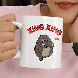 May include: White ceramic mug featuring the text "XING XING" in red letters. Below the text is a cartoon illustration of a smiling ape. The mug also has the Chinese characters for "stars".