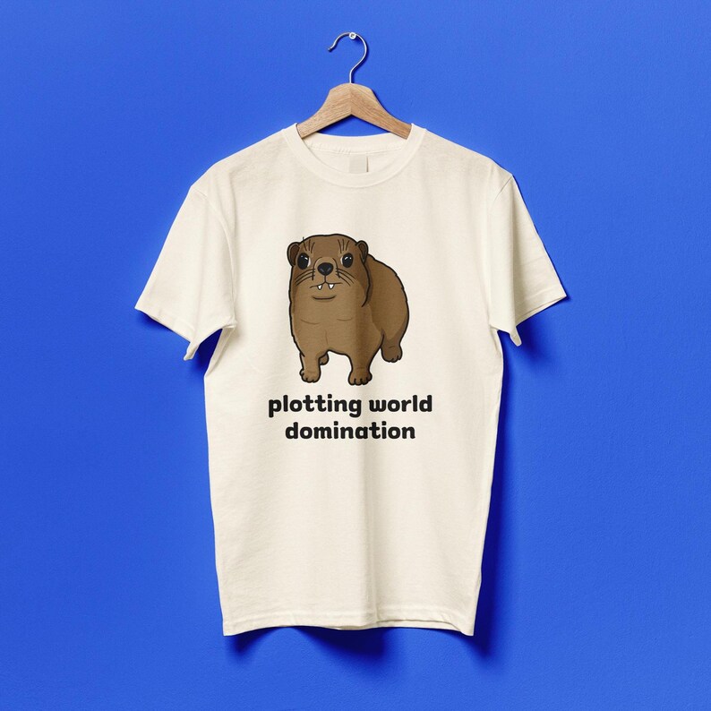 Cute brown hyrax illustration wearing a serious expression with the text “plotting world domination” printed on a t-shirt, funny animal meme graphic tee
