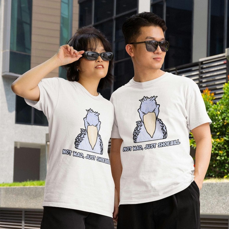 May include: A white short-sleeved t-shirt featuring a graphic illustration of a shoebill bird with a light blue body and a large beige beak. Below the bird, the text reads "NOT MAD, JUST SHOEBILL" in bold, dark blue capital letters.