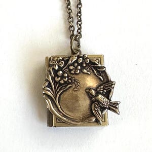 May include: A rectangular brass-toned metal locket pendant suspended from a matching link chain. The locket features a raised relief design on the front depicting a small bird perched near a cluster of flowers and leaves, with a smooth center.