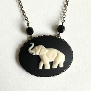 May include: An ivory-colored elephant pendant necklace with a black oval background. The elephant is raised and detailed. The necklace chain is a dark metal with black beads. A unique piece of jewelry.