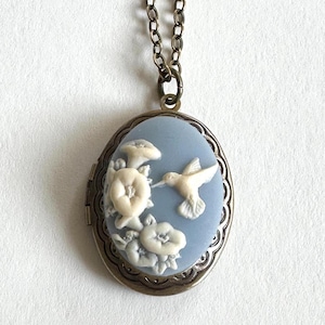 May include: A silver locket necklace with a blue enamel oval pendant featuring a white hummingbird and flowers.