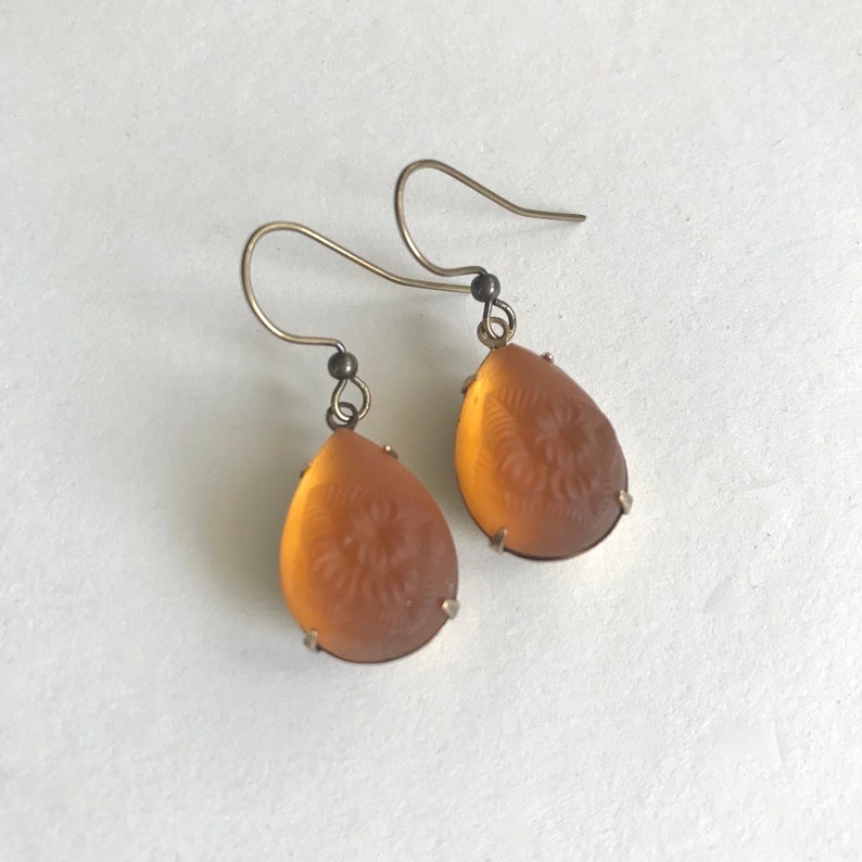 May include: A pair of vintage-style earrings. Each earring features a teardrop-shaped, amber-colored stone with an etched floral design. The stones are set in a silver-colored metal frame and hang from a simple hook.