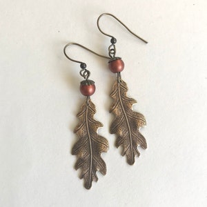May include: A pair of dangle earrings featuring a bronze oak leaf design with a textured surface and a small, round, copper-colored bead at the top of each earring.