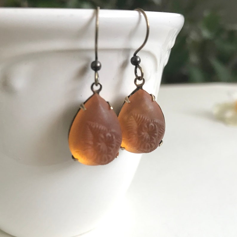 May include: A pair of teardrop-shaped earrings with a light amber hue. The earrings feature a detailed, etched design within the amber-colored glass, set in a dark metal frame with a hook closure. The earrings are displayed against a white background.