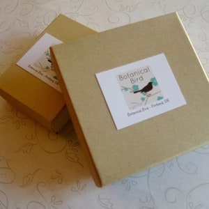 May include: Two square, light brown cardboard boxes with white labels. The labels feature a black bird perched on a branch with blue leaves and the text "Botanical Bird - Portland, OR". The boxes are slightly angled.