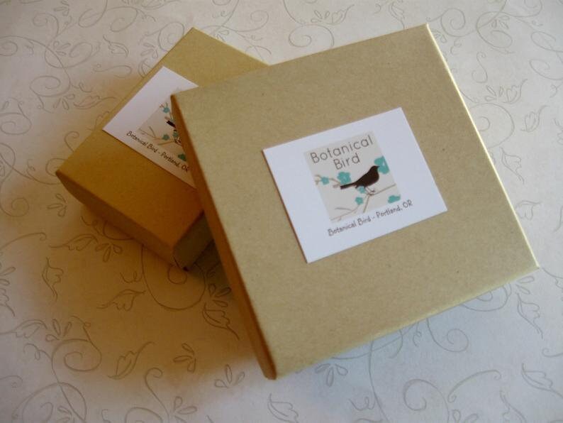 May include: Two square, light brown cardboard boxes with white labels. The labels feature a black bird perched on a branch with blue leaves and the text "Botanical Bird - Portland, OR". The boxes are slightly angled.
