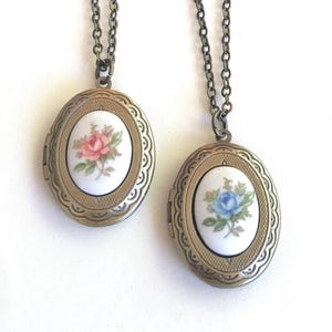 May include: Two oval-shaped metal lockets with antique bronze-toned finish and decorative scalloped edges. Each locket features a central white ceramic inlay; one displays a pink rose illustration, while the other shows a blue rose illustration.