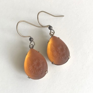 May include: A pair of teardrop-shaped earrings with a floral design etched into the amber-colored glass. The earrings have silver-colored metal hooks and settings, creating a vintage aesthetic.