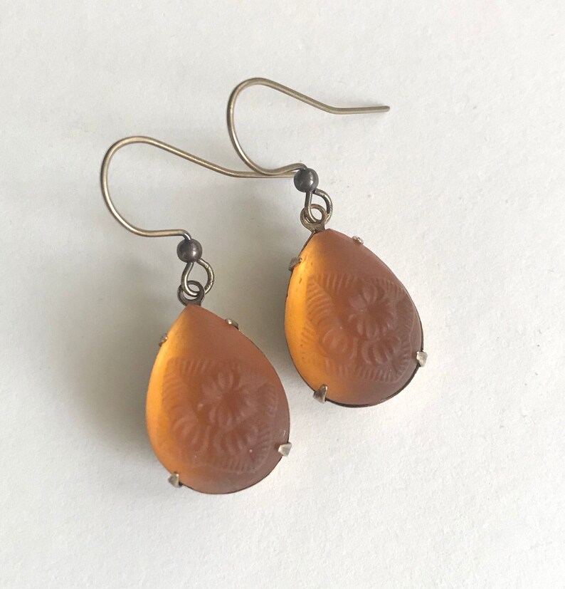 May include: A pair of teardrop-shaped earrings with a floral design etched into the amber-colored glass. The earrings have silver-colored metal hooks and settings, creating a vintage aesthetic.