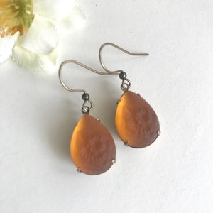 May include: A pair of teardrop-shaped earrings with a floral design etched into the amber-colored glass. The earrings have silver-colored hooks and are displayed on a white surface, with a white flower in the background.