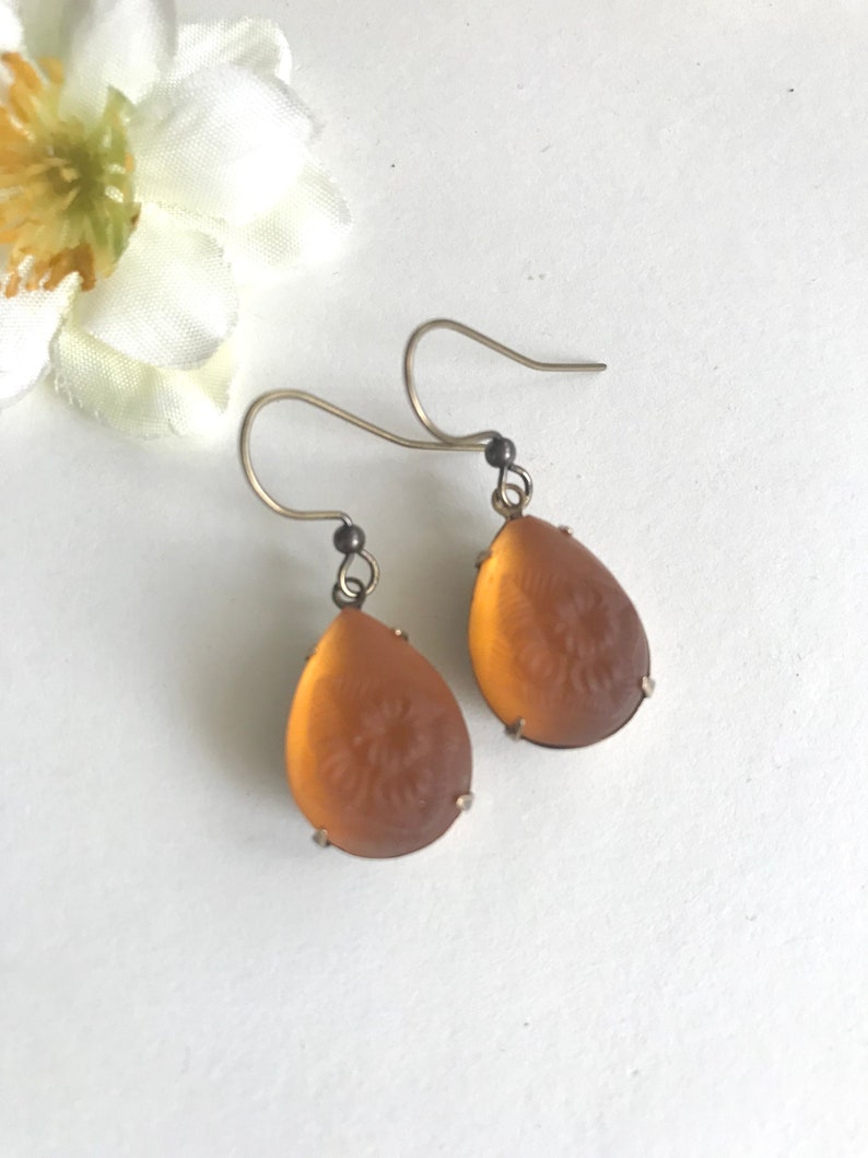 May include: A pair of teardrop-shaped earrings with a floral design etched into the amber-colored glass. The earrings have silver-colored hooks and are displayed on a white surface, with a white flower in the background.