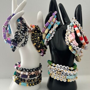 May include: A collection of colorful beaded bracelets with letter beads spelling out words and phrases like "FORTNITE", "ALL TOO WELL", "1989", and "READY".
