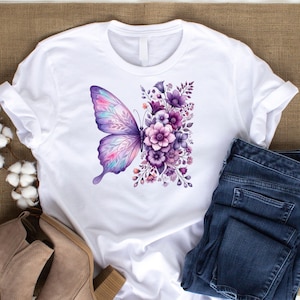 Puede incluir: Camiseta blanca con un dise&ntilde;o de mariposa en acuarela en tonos morados, rosas y azules. La mariposa est&aacute; colocada junto a un grupo de flores y follaje morado. La camiseta se muestra con vaqueros azules y botas color canela.