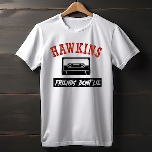 May include: White t-shirt with the word "HAWKINS" in red, above a black and white cassette tape graphic. Below the tape, the text "FRIENDS DON'T LIE" is printed in white on a black rectangle. The shirt is hanging on a wooden hanger.