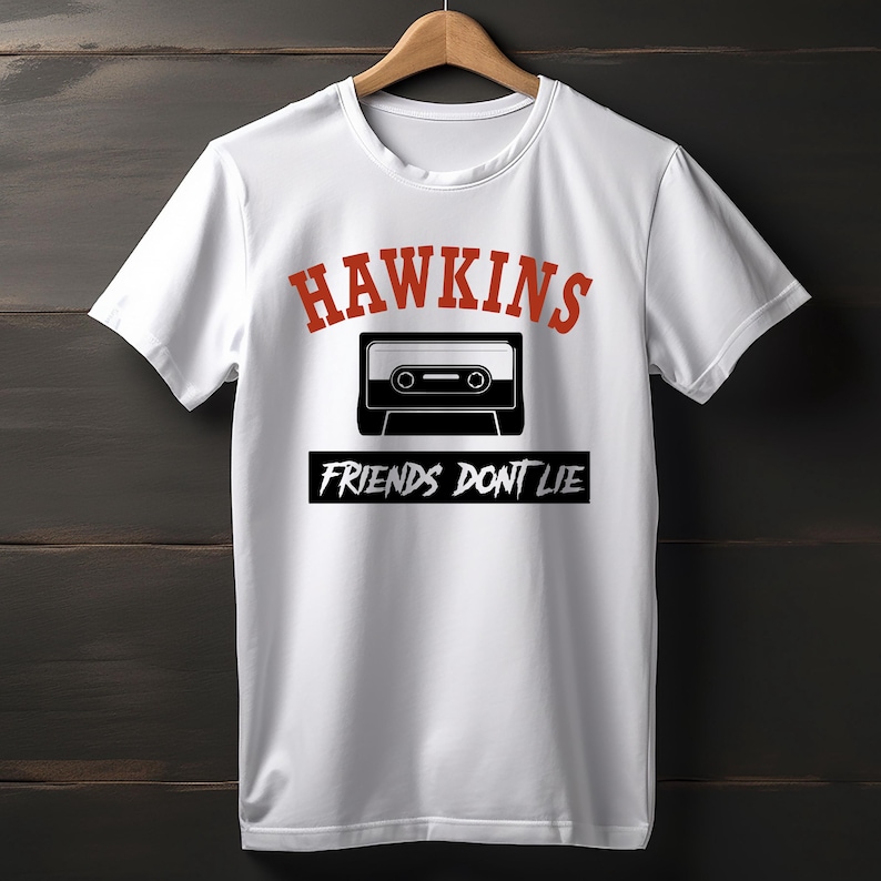 May include: White t-shirt with the word "HAWKINS" in red, above a black and white cassette tape graphic. Below the tape, the text "FRIENDS DON'T LIE" is printed in white on a black rectangle. The shirt is hanging on a wooden hanger.