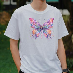 Puede incluir: Camiseta blanca con un dise&ntilde;o de mariposa colorida. La mariposa tiene alas rosas, moradas, azules y naranjas con patrones intrincados. La camiseta es de estilo cuello redondo.