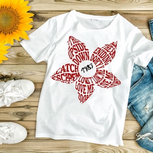 May include: White t-shirt with a red floral design. The flower design contains the words "Upside Down", "Catch", "Escape", "Love Me", and "Ending". The year "1983" is in the center of the flower.