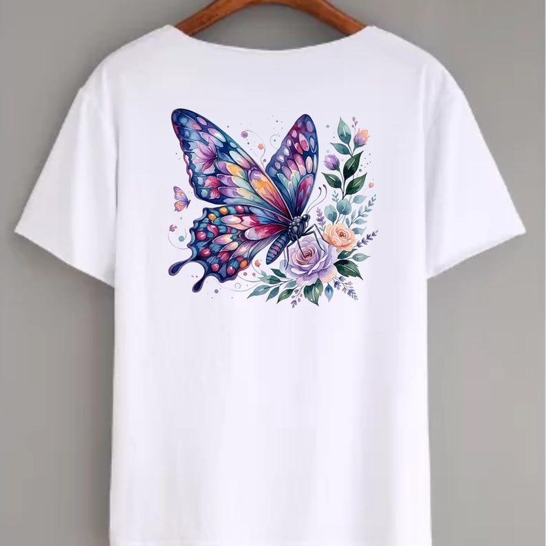 Puede incluir: Camiseta blanca con un dise&ntilde;o de mariposa en acuarela colorida. La mariposa tiene alas azules, moradas, rosas y naranjas, rodeada de flores y vegetaci&oacute;n. Tambi&eacute;n se ve una mariposa m&aacute;s peque&ntilde;a.