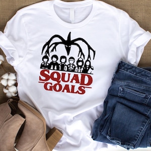 May include: White t-shirt with a black graphic of a creature and a group of people. Below the graphic, the words "SQUAD GOALS" are printed in red. The shirt is paired with blue jeans and tan boots.