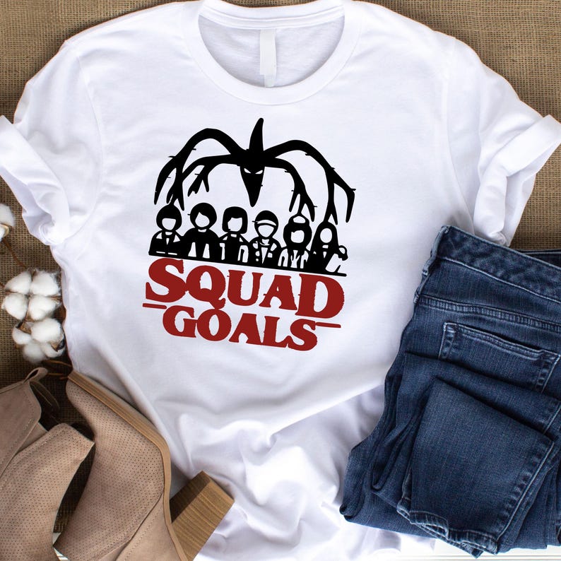 May include: White t-shirt with a black graphic of a creature and a group of people. Below the graphic, the words "SQUAD GOALS" are printed in red. The shirt is paired with blue jeans and tan boots.