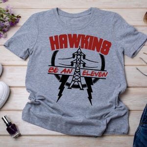 May include: A heather gray t-shirt featuring the text "HAWKINS" in red block letters above a black graphic of a power line tower and the words "BE AN ELEVEN". Two black lightning bolts are below the graphic.