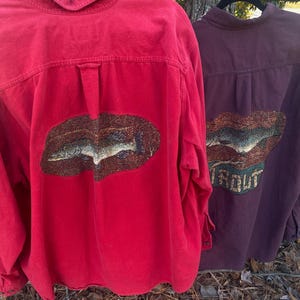 May include: Two button-down shirts hanging on black hangers. One is red with a fish design, the other is purple with a fish and the word "TROUT". The shirts are made of a soft material.
