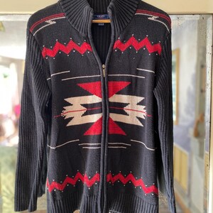 Könnte beinhalten: Ein schwarzer Reißverschluss-Pullover mit einem geometrischen Muster in Rot, Weiß und Beige. Der Pullover hat einen hohen Kragen und gerippte Bündchen und Saum.