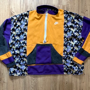 Könnte beinhalten: Ein mehrfarbiger Pullover mit einem halben Reißverschluss. Der Pullover besteht aus einem Patchwork aus verschiedenen Stoffen, darunter ein Blumendruck, einfarbiges Lila und ein gelber Stoff mit dem Nike-Logo. Der Pullover hat eine große Tasche auf der Vorderseite.