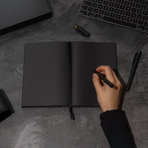 May include: A black leather-bound notebook with a black ribbon bookmark. A person is writing in the notebook with a black pen.
