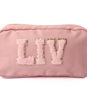 May include: A pale pink cosmetic bag featuring the name "LIV". The letters are a light pink, fluffy material with gold glitter edging. The bag has a zip closure.