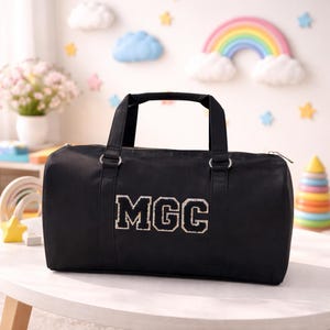 May include: Black duffel bag with the letters "MGC" in a white and silver design. The bag has two black handles and a zip closure. The bag is sitting on a white table.