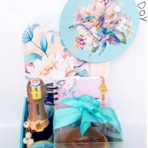 Puede incluir: Una cesta de regalo con un tema azul y floral, que contiene una botella de champán, un pastel y un cuaderno. La cesta está decorada con una cinta verde azulado y una tarjeta redonda con una ilustración de colibrí. Las palabras "Happy Mother Day" están escritas en la parte superior.