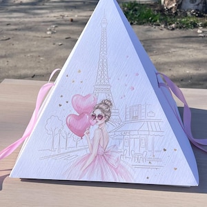 Puede incluir: Una caja de regalo blanca en forma de pirámide con una cinta rosa. La caja presenta una ilustración de la Torre Eiffel, una mujer sosteniendo globos en forma de corazón y una escena de café parisino. Los acentos rosas y los corazones dorados añaden un toque romántico.