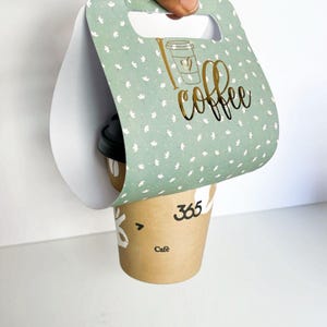Peut inclure: Un porte-gobelet en papier vert et blanc avec un motif "J'aime le café" en or. Le porte-gobelet a une poignée et est conçu pour contenir un gobelet à café de taille standard.