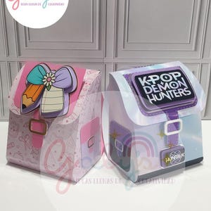 Puede incluir: Dos cajas decorativas de papel con forma de mochilas. Una es rosa con un diseño de lápiz y lazo, la otra es blanca y morada con el texto "KPOP DEMON HUNTERS". Ambas tienen hebillas y están sobre una superficie blanca.