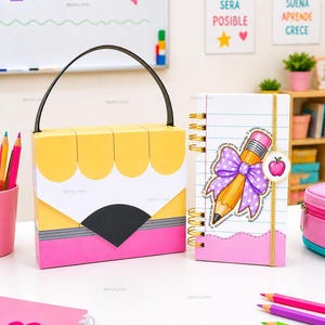 Könnte beinhalten: Eine gelb-rosa stiftförmige Box mit schwarzem Griff und ein Spiralnotizbuch mit Bleistift- und Apfeldesign. Das Notizbuch hat einen rosa Einband und einen goldenen Gummibandverschluss. Buntstifte befinden sich in einem rosa Becher.