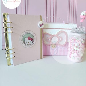 May include: A light pink ring-bound planner with a Hello Kitty shaker charm, a white and pink rectangular storage box featuring a glittery bow accent, and a clear glass tumbler with a pink lid, straw, and Hello Kitty character graphics.