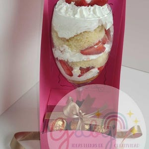以下が含まれることがあります： ホイップクリームとイチゴを添えた、背の高いグラスに入った層状のデザートが入ったピンク色のギフトボックス。箱には、リボンで結ばれたチョコレートが入った小さな箱も入っています。「IDEAS LLENAS DE CREATIVIDAD」の文字が見えます。