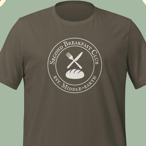 Op de afbeelding: Olijfgroen T-shirt met een wit cirkelvormig logo. Het logo toont een vork en mes gekruist over een brood, met de tekst "SECOND BREAKFAST CLUB" en "EST. MIDDLE-EARTH" in een cirkel.