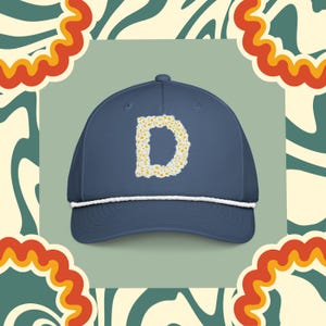 Op de afbeelding: Een marineblauwe baseballcap met een wit touwdetail langs de rand. De letter "D" is op de voorkant geborduurd met een bloemenontwerp in geel en wit. De cap heeft een gebogen klep en een klassieke, casual stijl.