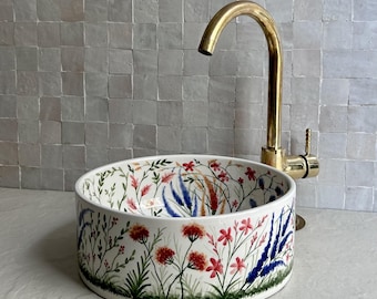 Puede incluir: Un lavabo de cerámica blanca con un diseño floral con flores rojas, rosas y azules, y follaje verde. Un grifo dorado está instalado sobre el lavabo. El lavabo es una cuenca redonda y poco profunda.