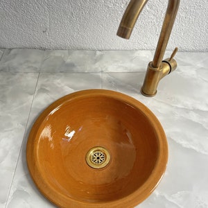 Pode incluir: Uma pia de terracota com uma torneira de latão. A pia é redonda e tem um ralo no centro. A torneira é de uma só alavanca com um bico curvo.