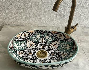 May include: Oval ceramic sink with a colorful Moroccan-inspired design. The sink features intricate floral patterns in shades of green, blue, black, and white. A brass faucet is visible in the background.